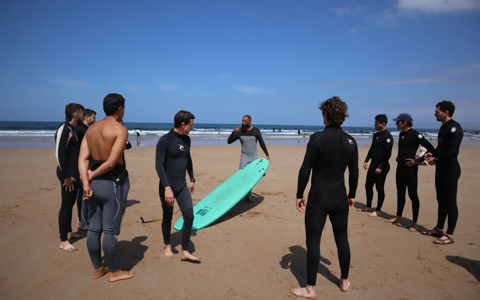 Surf group gathered around a turquoise board for a beach briefing — previous MatnWave edition